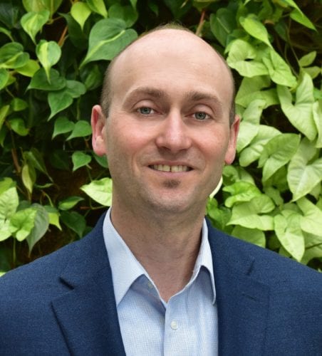 Man in a blue suit smiling outdoors with green foliage background.