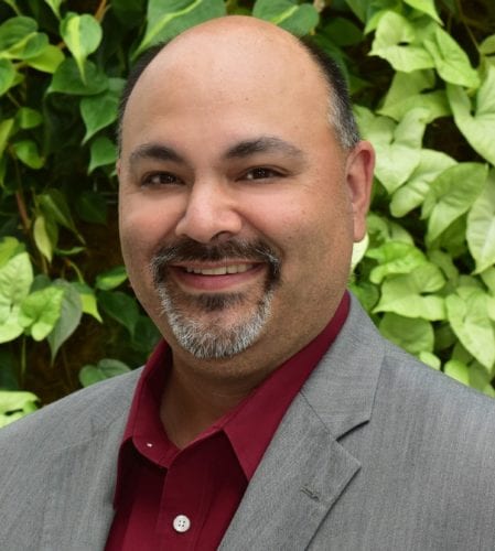 Smiling man in a gray suit with green foliage background.