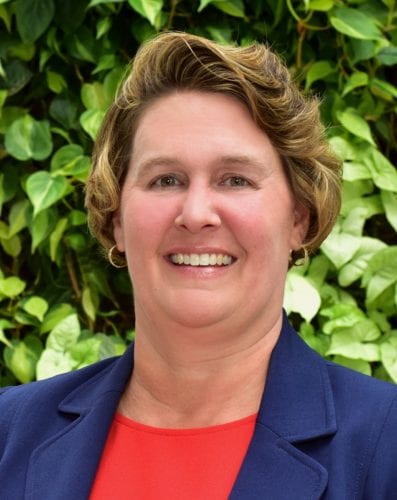 Smiling woman in a blue blazer with greenery background.