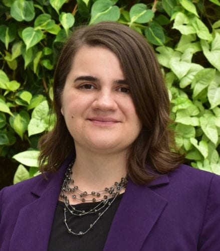 Smiling woman in a purple blazer standing outdoors with green foliage behind her.