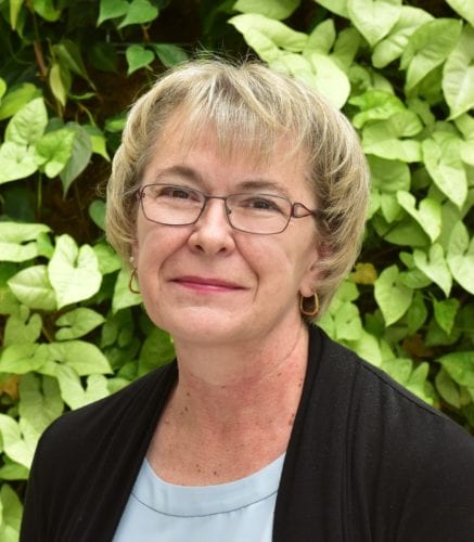 Smiling woman with glasses in front of green foliage.