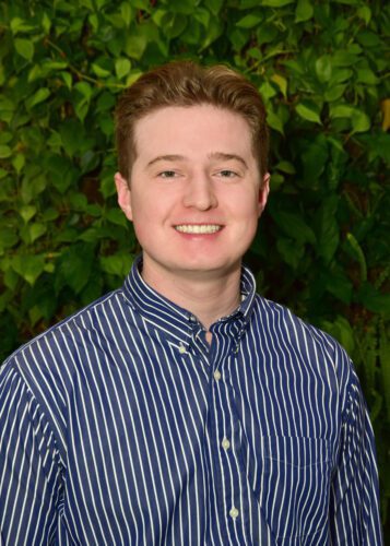 A young man smiling in front of leafy green background.