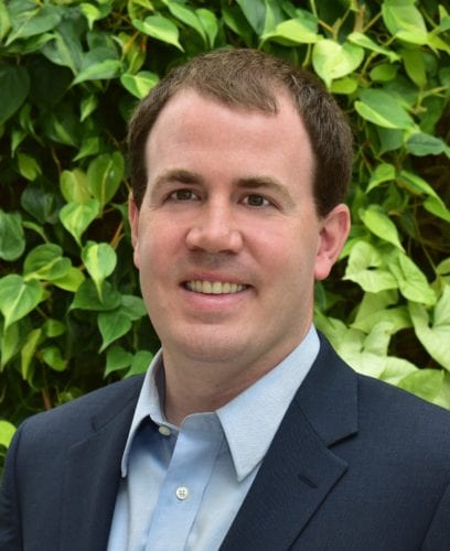 Man in a suit smiling outdoors with green foliage background.
