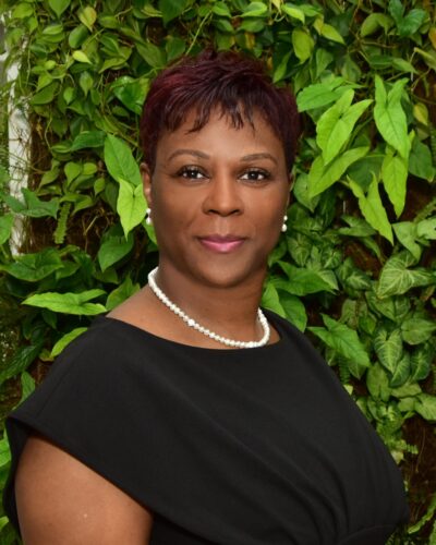 Professional woman in a black dress with a green leafy background.