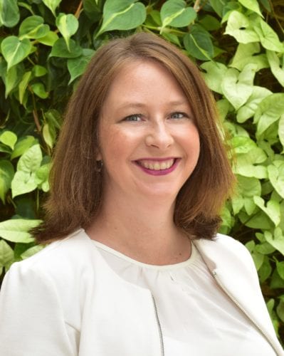 Smiling woman with shoulder-length hair in a white blouse stands before green foliage.
