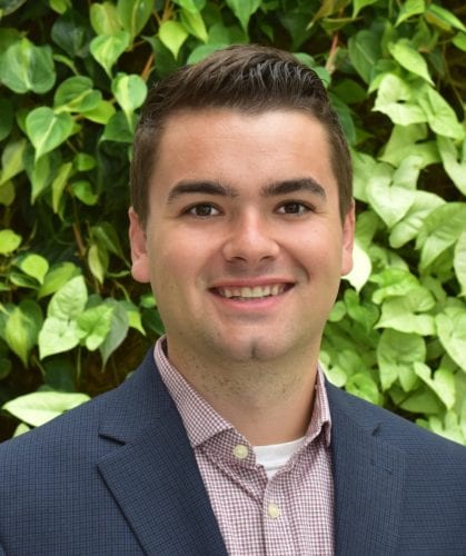 Young man in a suit smiling outdoors with green leafy background.