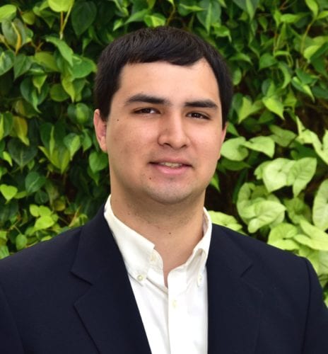 A man in a suit stands before a lush green background.