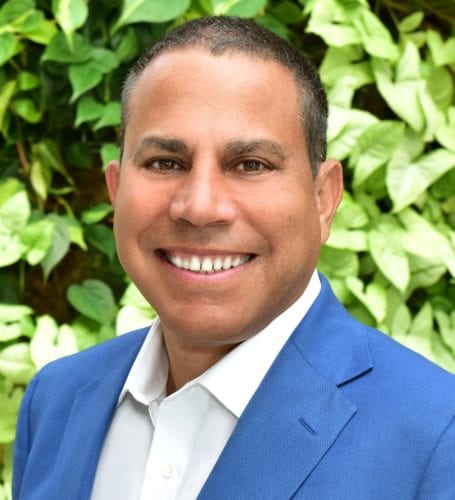 Smiling man in a blue suit with a leafy background.