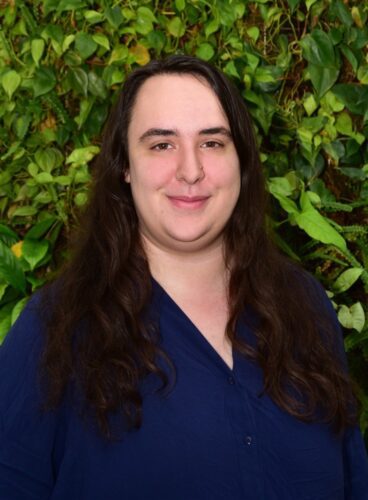 A person smiling with long dark hair against a leafy green background.
