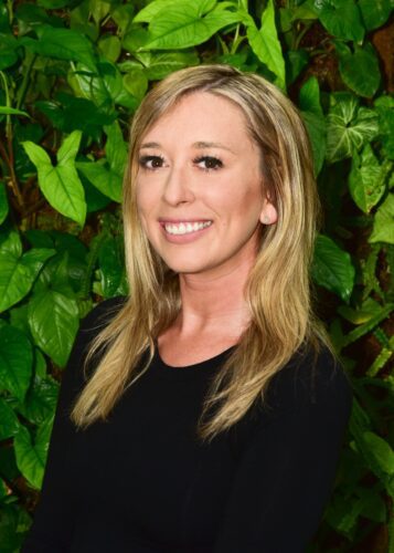 Smiling woman with blonde hair in front of green foliage.