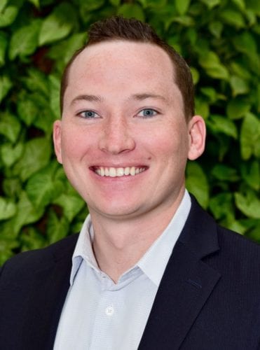 A smiling man in a suit with a leafy green background.