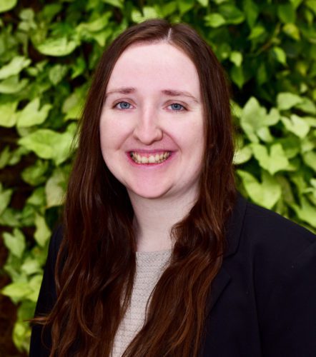 A woman with long brown hair smiling in front of green foliage.