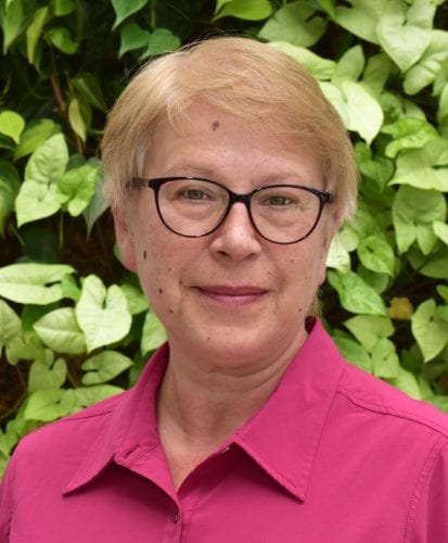 Portrait of a smiling woman with glasses and short blonde hair in front of green foliage.