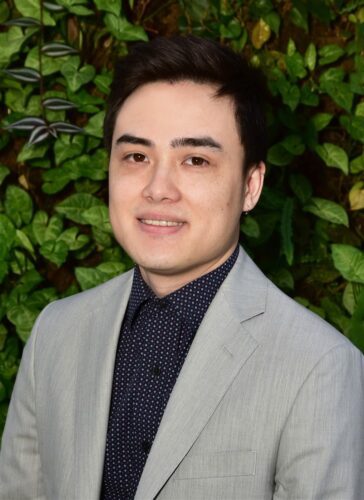 Young man in a gray blazer smiling against a leafy background.