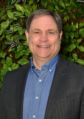 Smiling man in a blue shirt and dark blazer stands before green foliage.