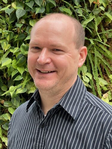 Smiling man in a striped shirt with green foliage background.