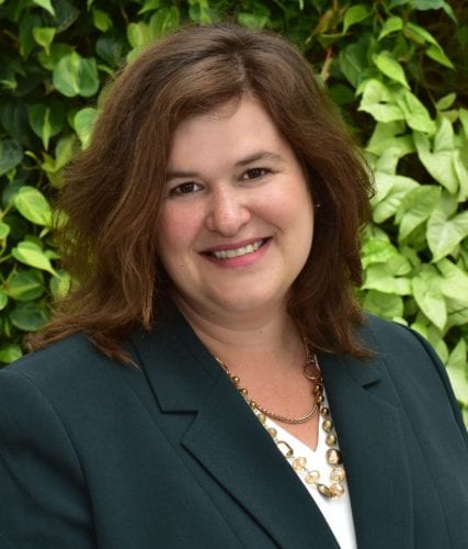 Smiling woman in a dark blazer with green leafy background.