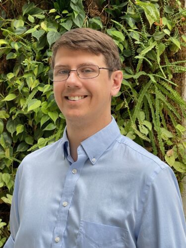 Smiling man wearing glasses and a blue button-up shirt outdoors.