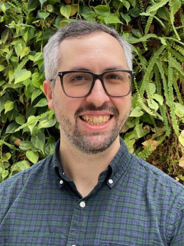 Smiling man with glasses in front of green foliage.