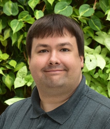 Smiling man with short brown hair in front of green leafy background.