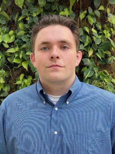 A young man in a blue button-up shirt stands in front of green foliage.