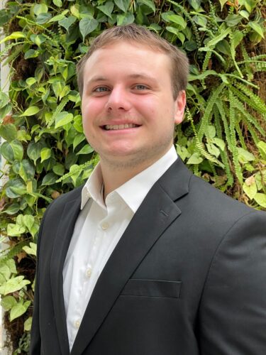 Young man in a black suit smiling outdoors with greenery background.