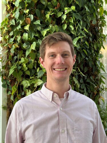 Smiling man in a light shirt stands before a green leafy background.