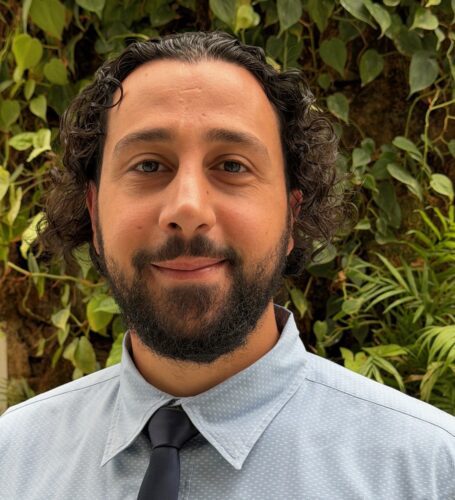 A man with curly hair and a beard wearing a light blue shirt and tie.