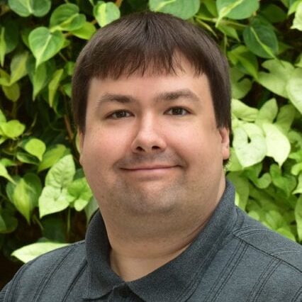 Smiling man with short brown hair in front of green leafy background.