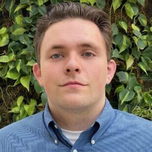 A young man in a blue button-up shirt stands in front of green foliage.