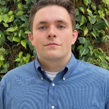 A young man in a blue button-up shirt stands in front of green foliage.