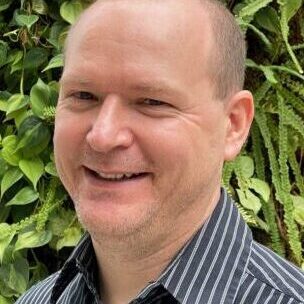 Smiling man in a striped shirt with green foliage background.