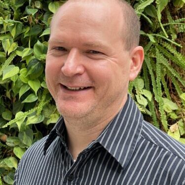 Smiling man in a striped shirt with green foliage background.