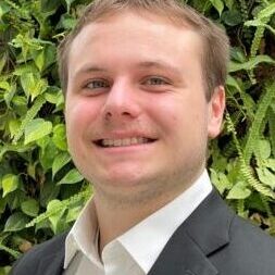 Young man in a black suit smiling outdoors with greenery background.
