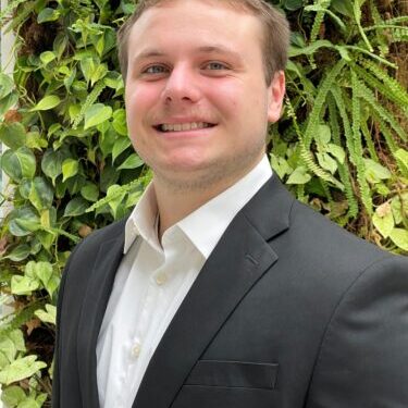 Young man in a black suit smiling outdoors with greenery background.