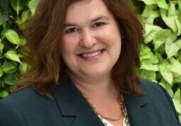Smiling woman in a dark blazer with green leafy background.