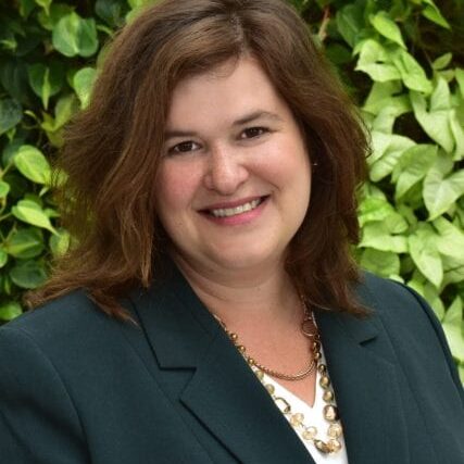 Smiling woman in a dark blazer with green leafy background.