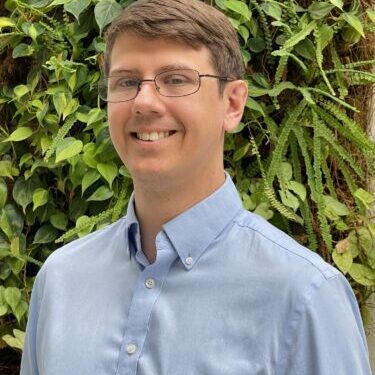 Smiling man wearing glasses and a blue button-up shirt outdoors.