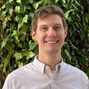 Smiling man in a light shirt stands before a green leafy background.