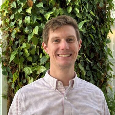 Smiling man in a light shirt stands before a green leafy background.