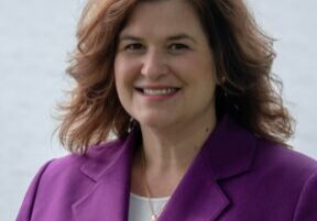 A smiling woman wearing a purple blazer outdoors by the water.