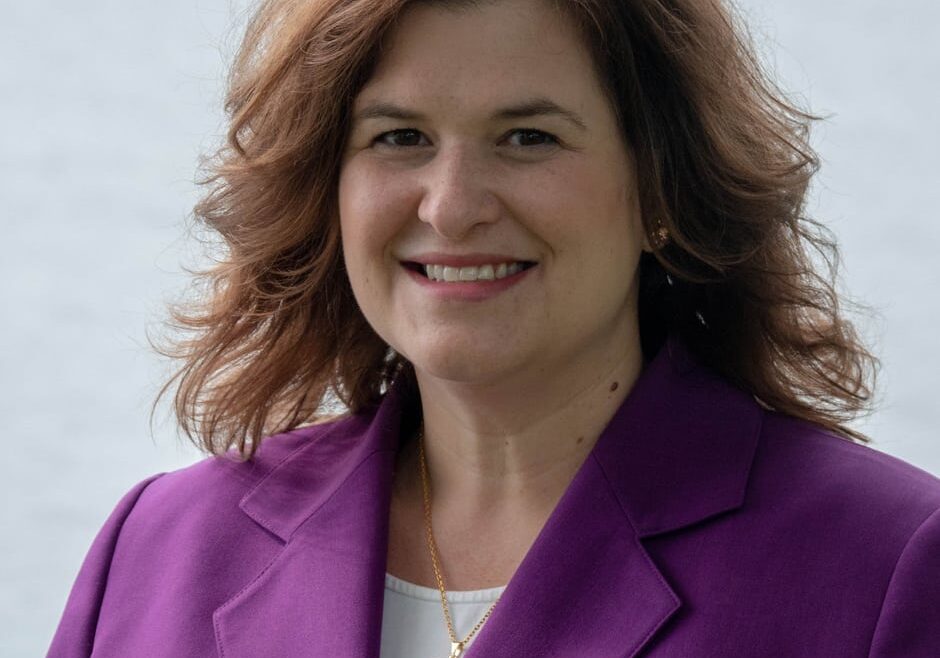 A smiling woman wearing a purple blazer outdoors by the water.
