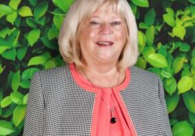 Smiling woman with blonde hair, wearing a gray jacket and coral blouse, standing against a leafy green background.