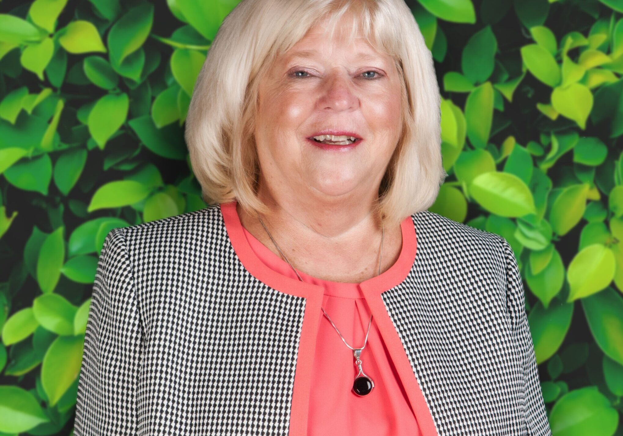 Smiling woman with blonde hair, wearing a gray jacket and coral blouse, standing against a leafy green background.