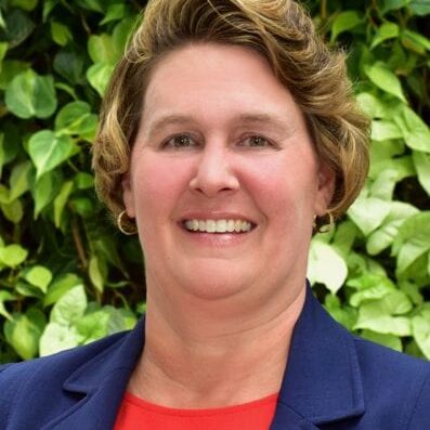 Smiling woman in a blue blazer with greenery background.