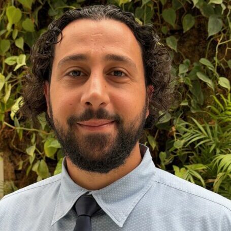 A man with curly hair and a beard wearing a light blue shirt and tie.