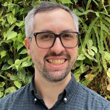 Smiling man with glasses in front of green foliage.