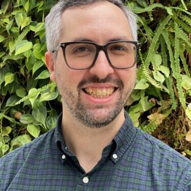 Smiling man with glasses in front of green foliage.