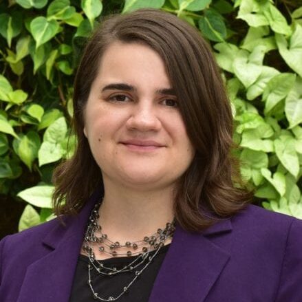 Smiling woman in a purple blazer standing outdoors with green foliage behind her.