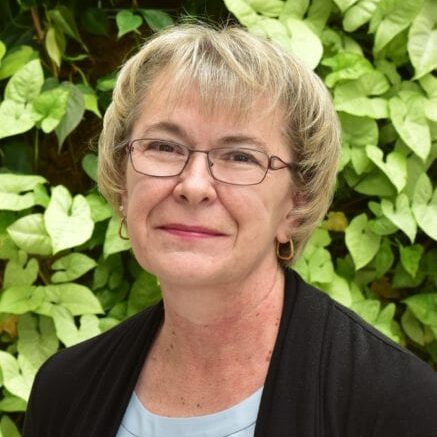 Smiling woman with glasses in front of green foliage.